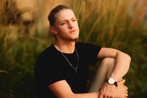 Young man with tied-back hair wearing black t-shirt and watch sitting in golden sunlit grass field.