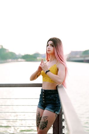 Young woman with pink ombre hair in yellow crop top and denim shorts posing by waterfront.