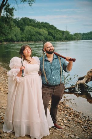 Couple standing by riverside, woman in white gown holding flute, man in teal shirt with axe.