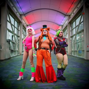 Three women in vibrant cosplay costumes pose together in colorful convention center hallway.