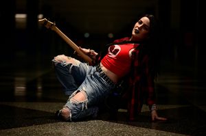 Woman in red crop top and ripped jeans playing guitar while sitting on floor dramatically.