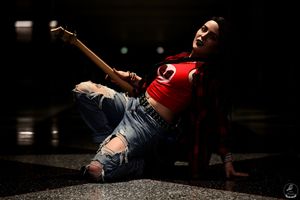Woman in red crop top and ripped jeans poses dramatically on floor with guitar.
