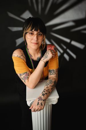 Tattooed woman with short hair and glasses leans on white pedestal against dark background.