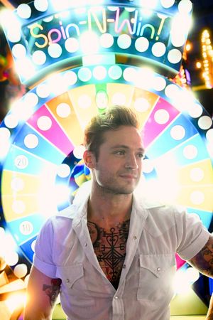 Tattooed man in white shirt stands before colorful Spin-N-Win carnival wheel at night.