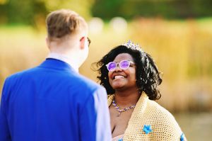 Smiling bride wearing tiara and purple glasses looks at groom during outdoor wedding.
