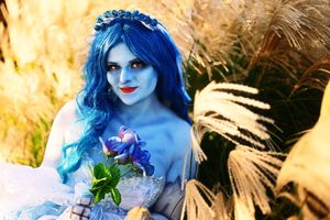 Woman cosplaying as the Corpse Bride with blue skin, blue wig, and floral bouquet in golden grass.