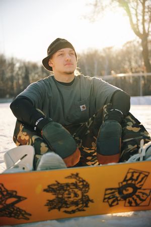 Young male snowboarder sitting in snow wearing Carhartt beanie and camouflage pants with orange snowboard.