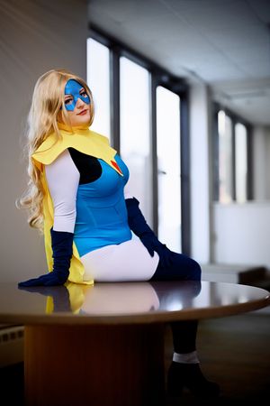 Blonde woman in blue and yellow superhero cosplay costume sitting on a table indoors.