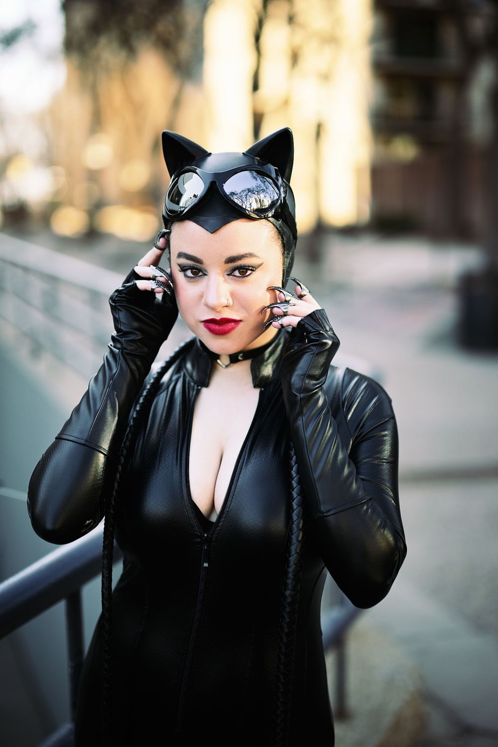 Woman in black Catwoman cosplay costume with cat ears and goggles posing outdoors.