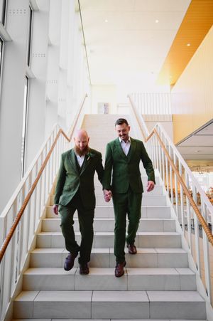 Two men in green suits holding hands while walking down modern interior staircase.
