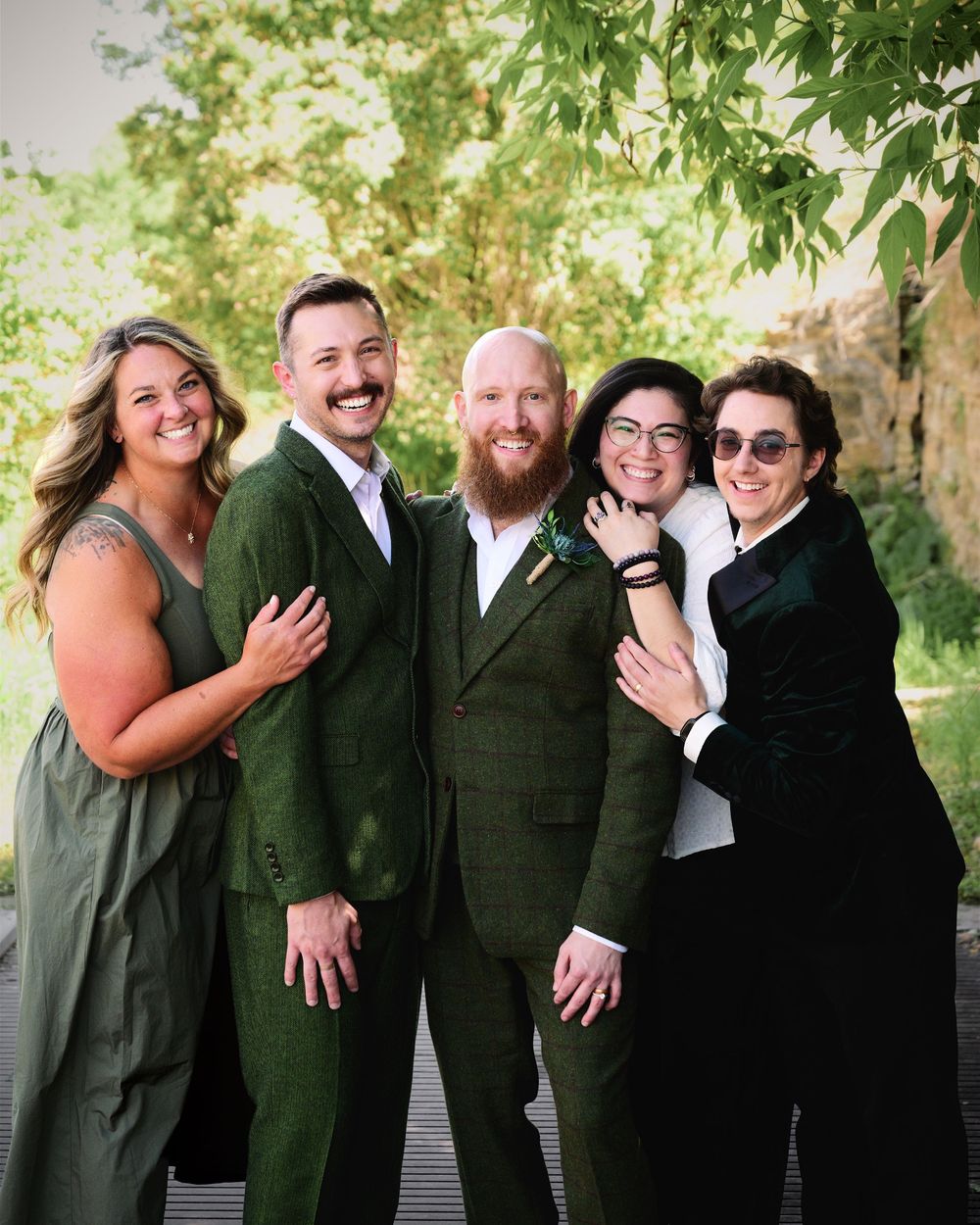 Group of five people smiling together outdoors, two wearing matching green suits at a wedding.