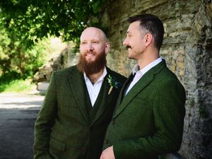 Two men in matching green tweed suits smiling together against a stone wall outdoors.