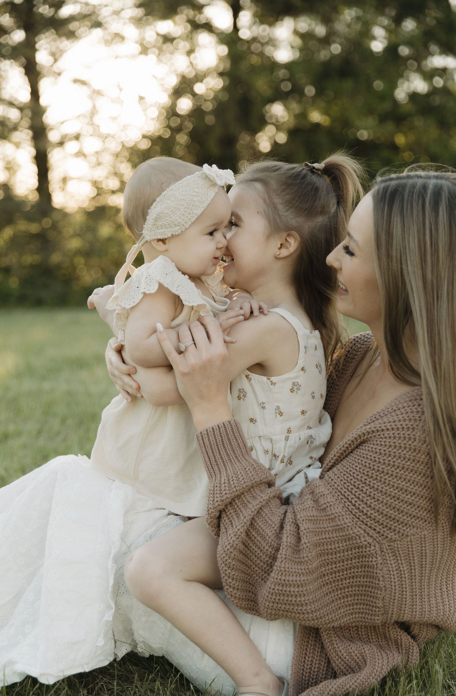 Mother in white dress holds baby while young daughter walks beside her along white farm fence at sunset.