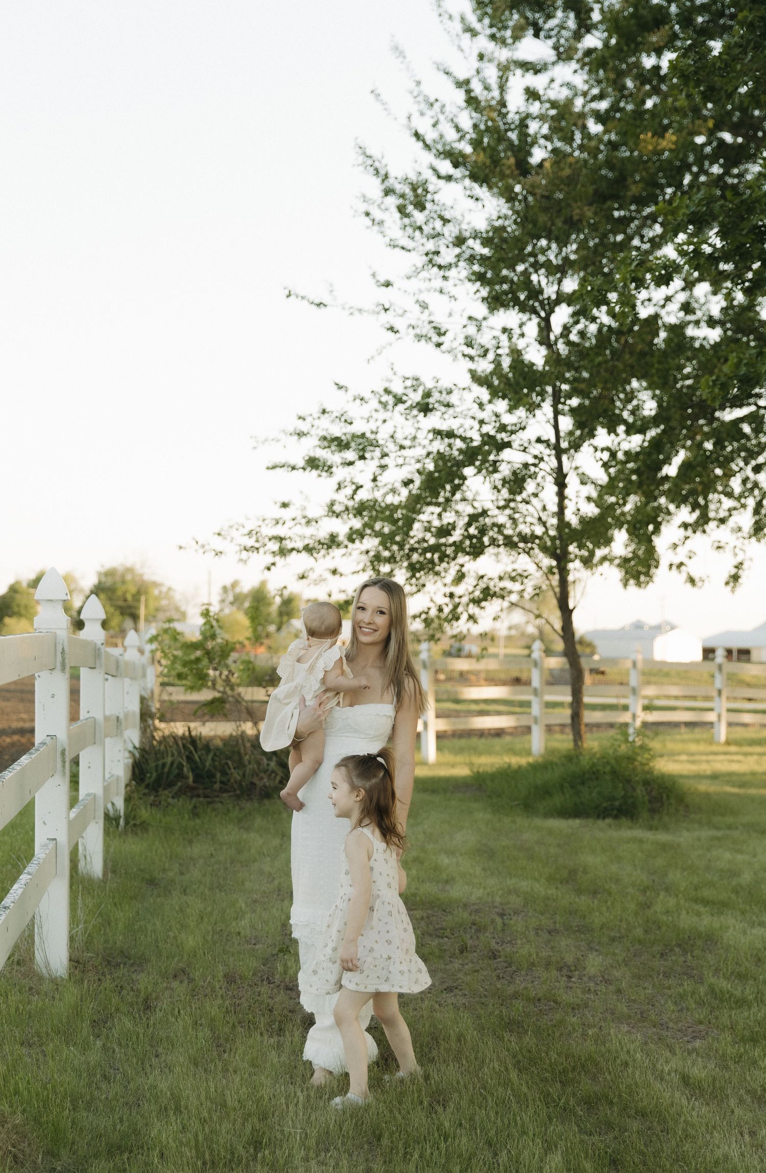 Mother in white dress holds baby while young daughter walks beside her along white farm fence at sunset.