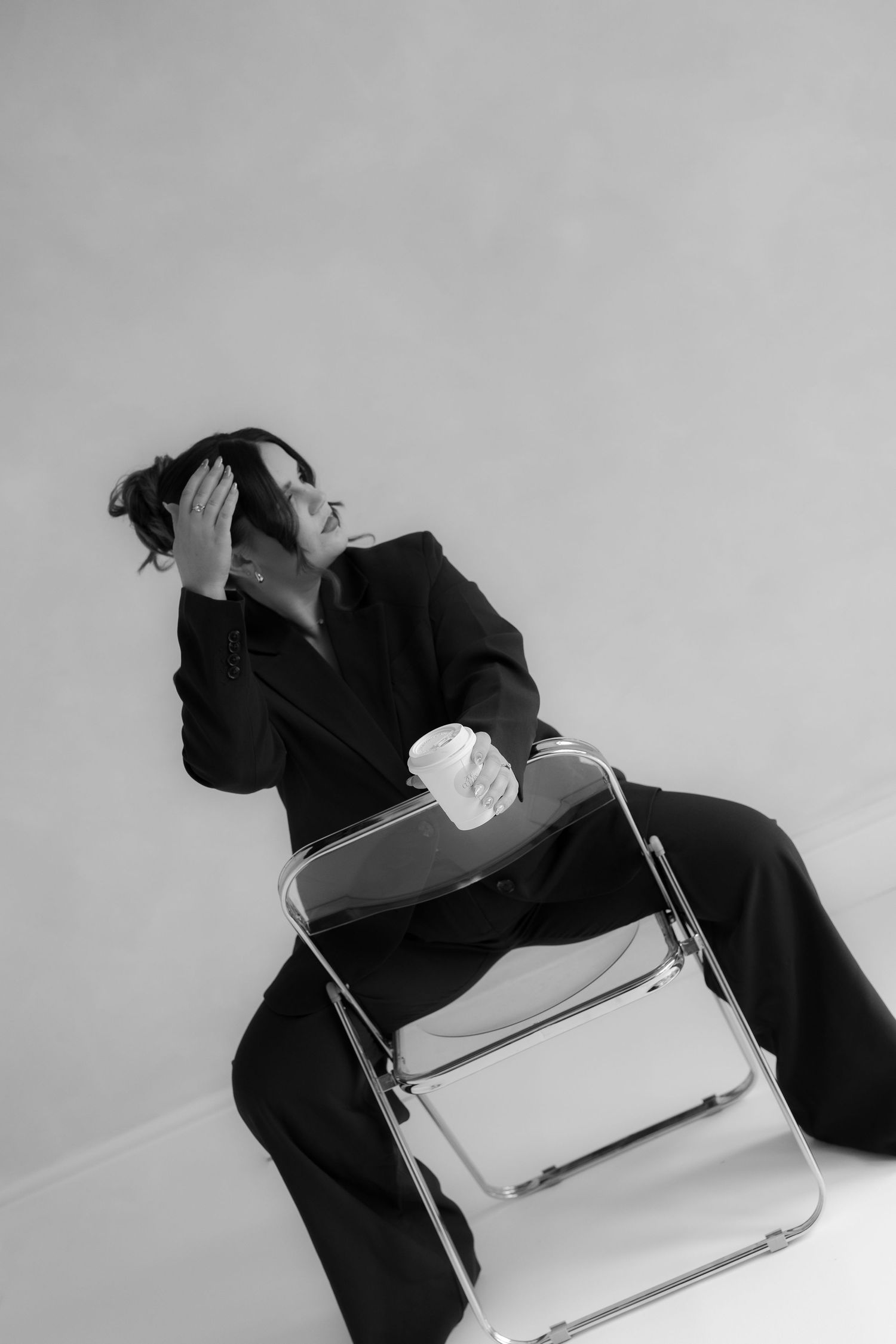 black and white image of photographer branding session while sitting backwards on a clear chair holding a coffee playing with her hair.