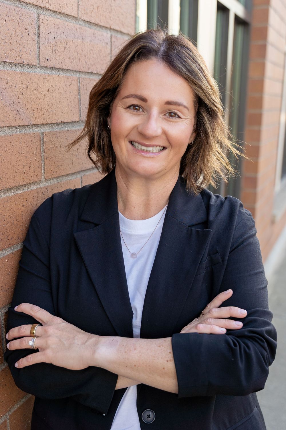 Professional portrait of someone in a black blazer and white shirt smiling while leaning against a brick wall.