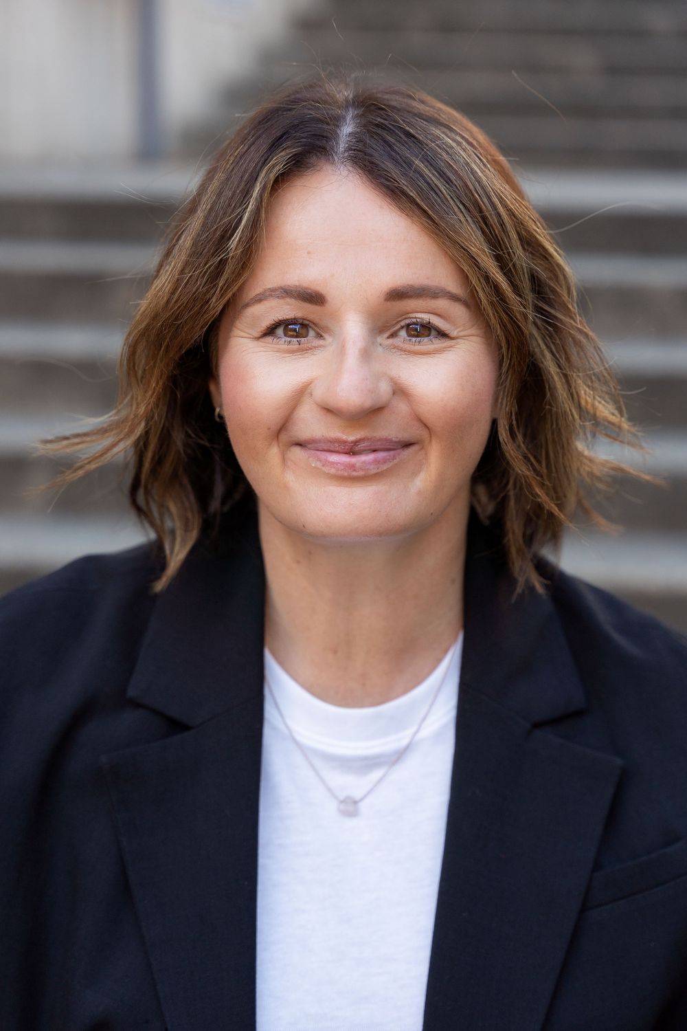Professional headshot series showing different expressions while wearing a black blazer and white top against blurred background.