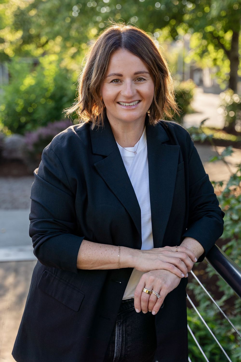 Professional portrait of someone in a black blazer and white shirt sitting outdoors with natural lighting and bokeh background.