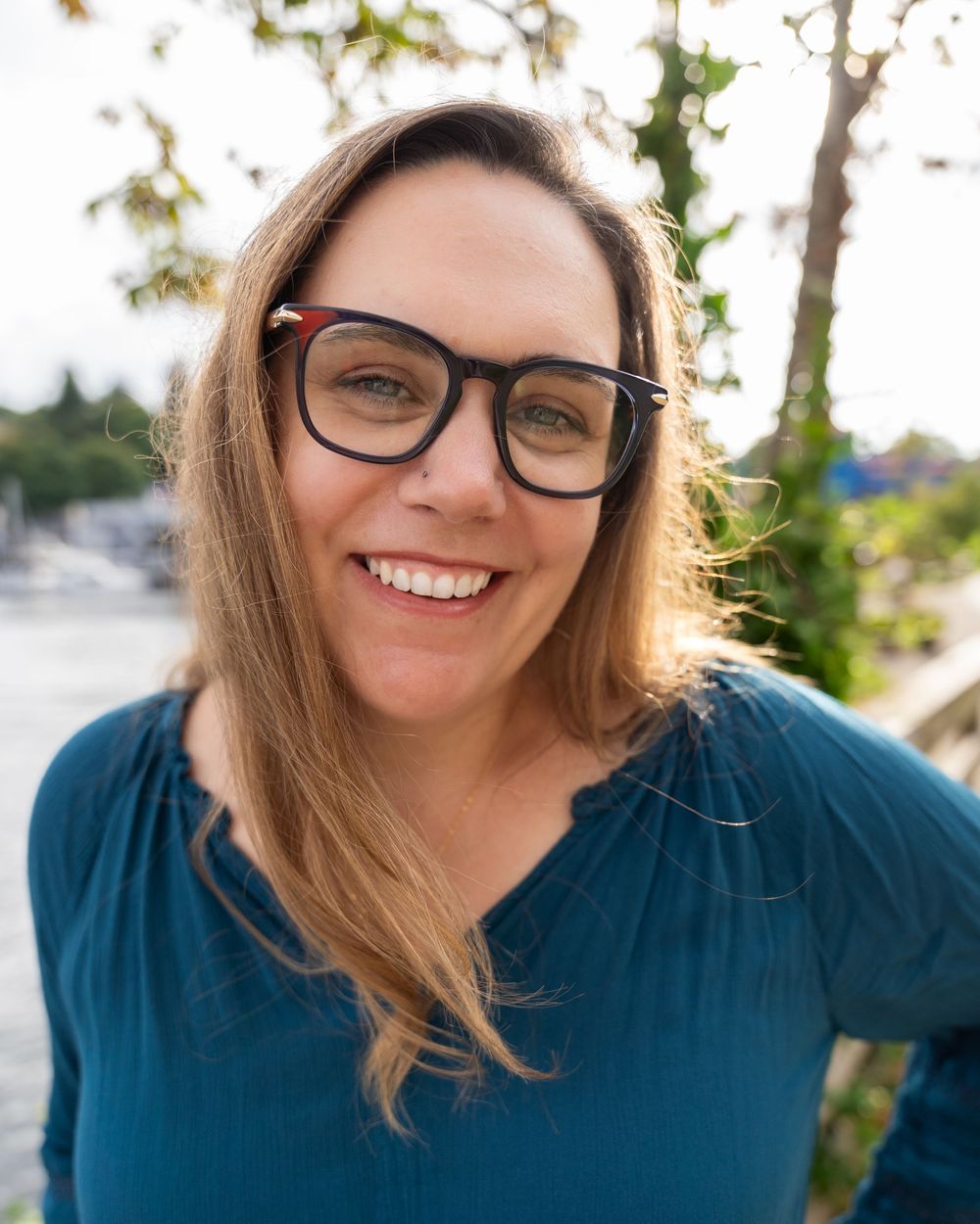Person with glasses and long hair smiles warmly while wearing a navy blue top in outdoor setting.
