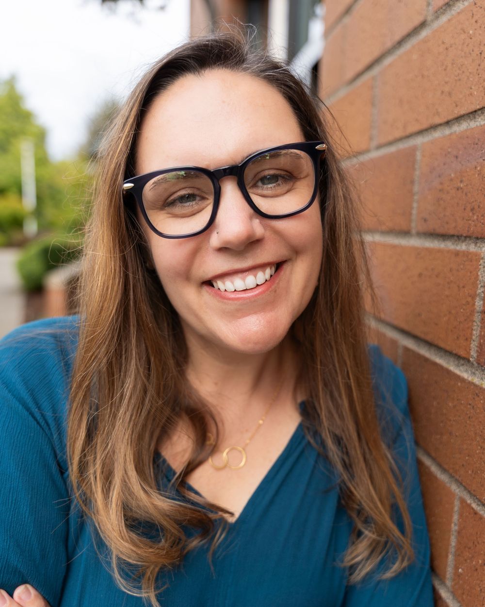 Professional headshots of someone in glasses and a blue top smiling against a brick wall background.