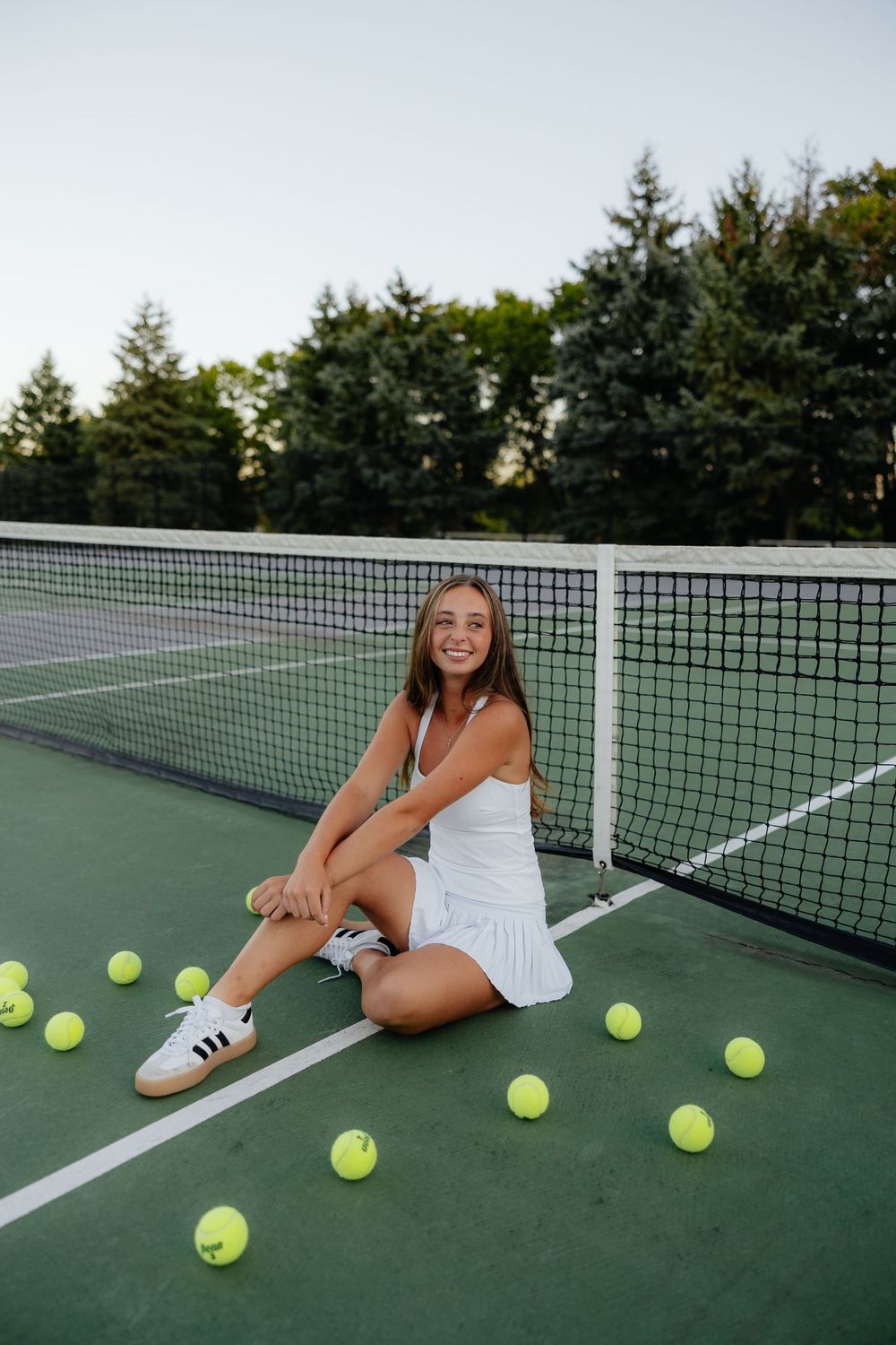 Tennis player in white dress sits on court surrounded by yellow tennis balls.