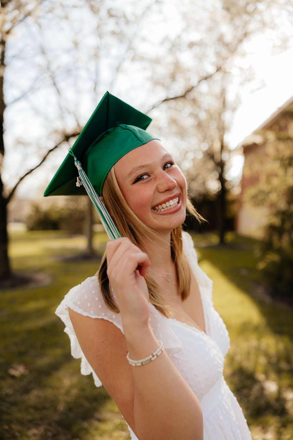 Graduate in white dress and green cap smiling outdoors in spring.