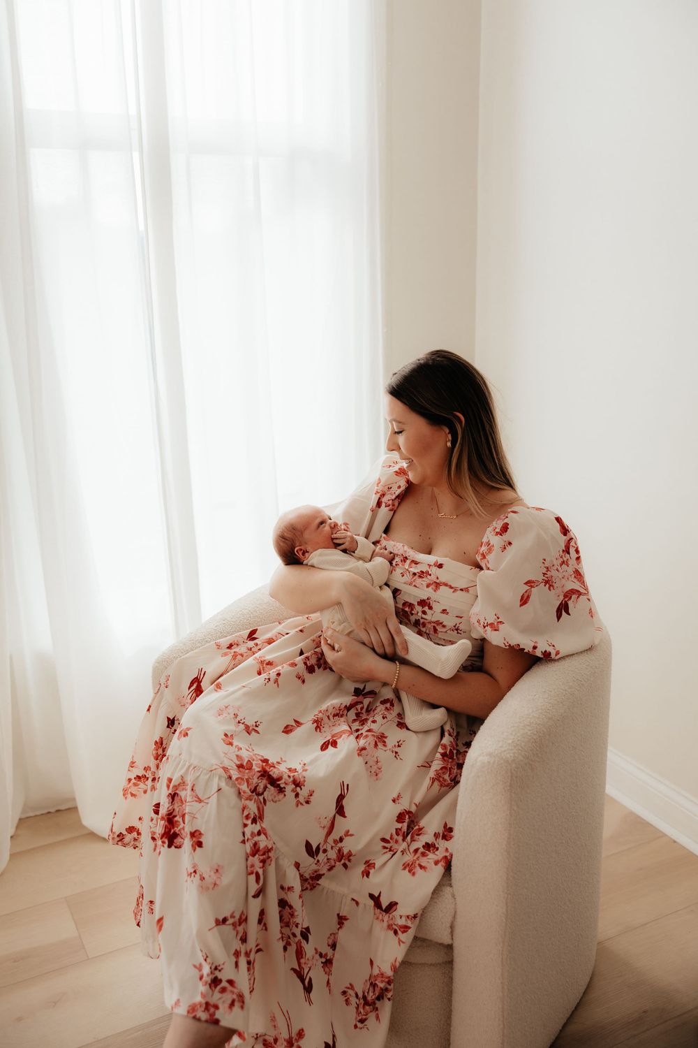 Mother in floral dress cradles newborn baby in white chair by window.