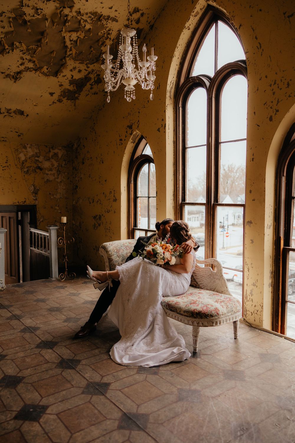 Wedding couple shares intimate moment on vintage sofa beneath arched windows and chandelier.