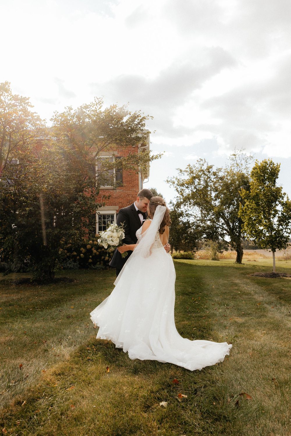 A romantic wedding photo of a couple embracing outside a rustic house on a cloudy day.