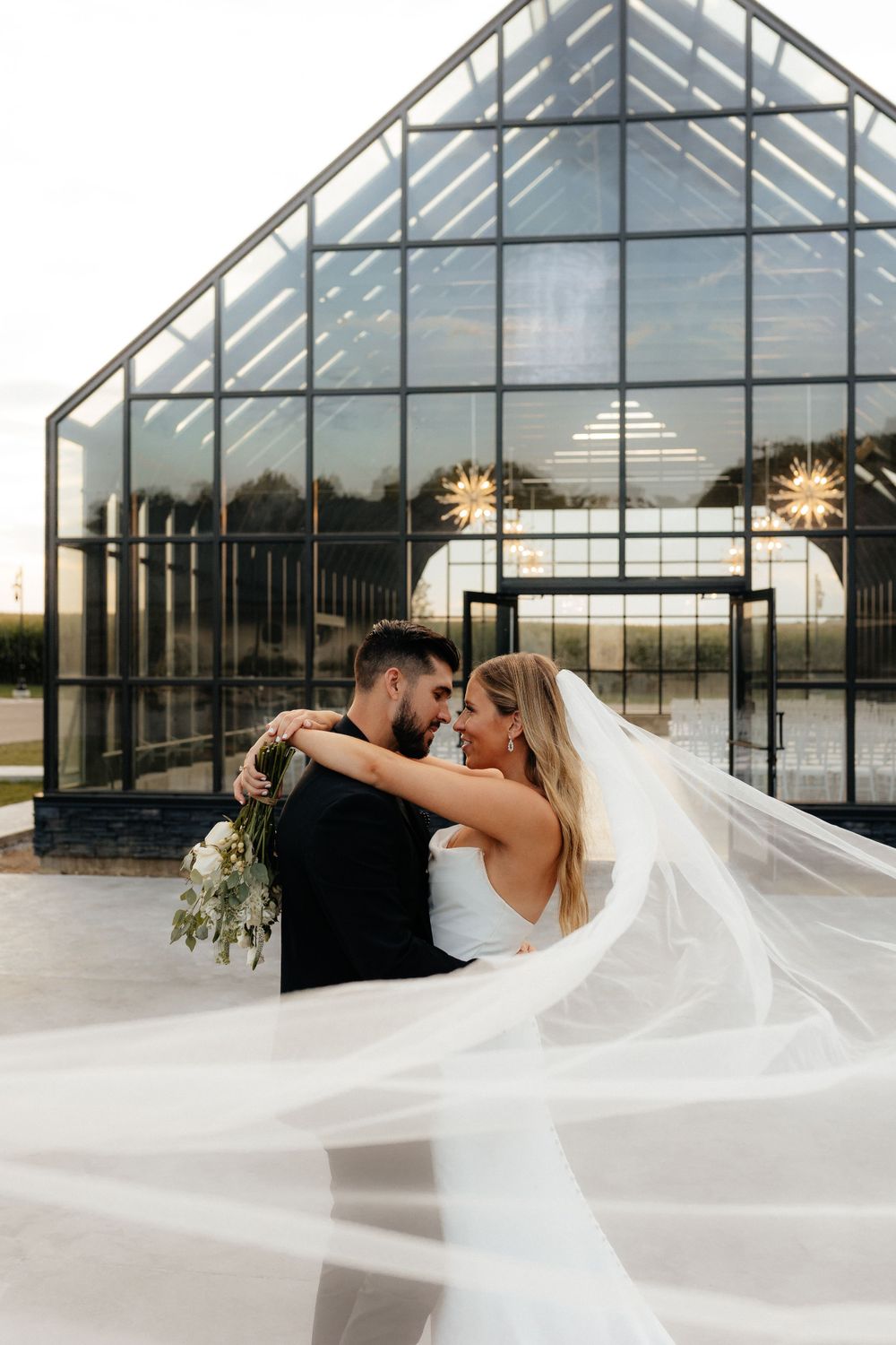 Romantic wedding couple embraces in front of a modern glass greenhouse with flowing white veil.