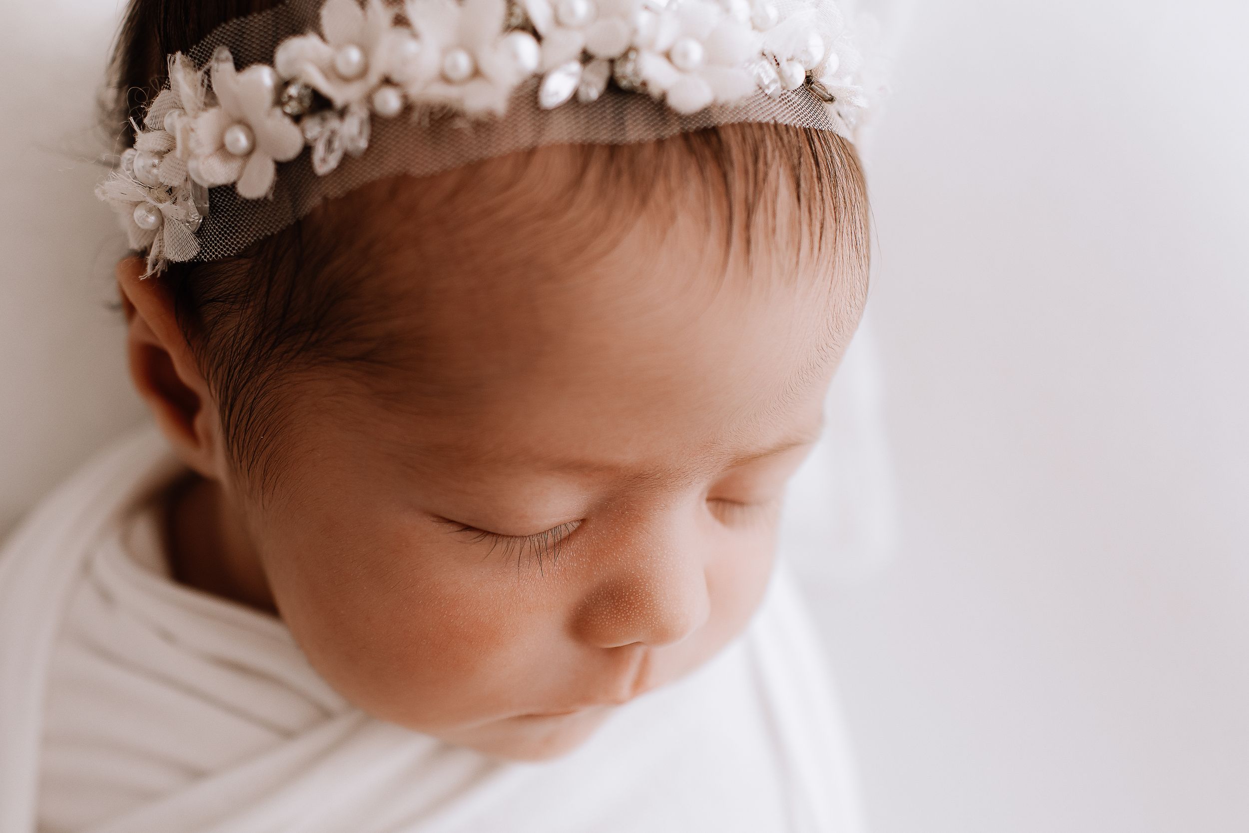 Close-up newborn portrait with delicate white floral crown against light background.
