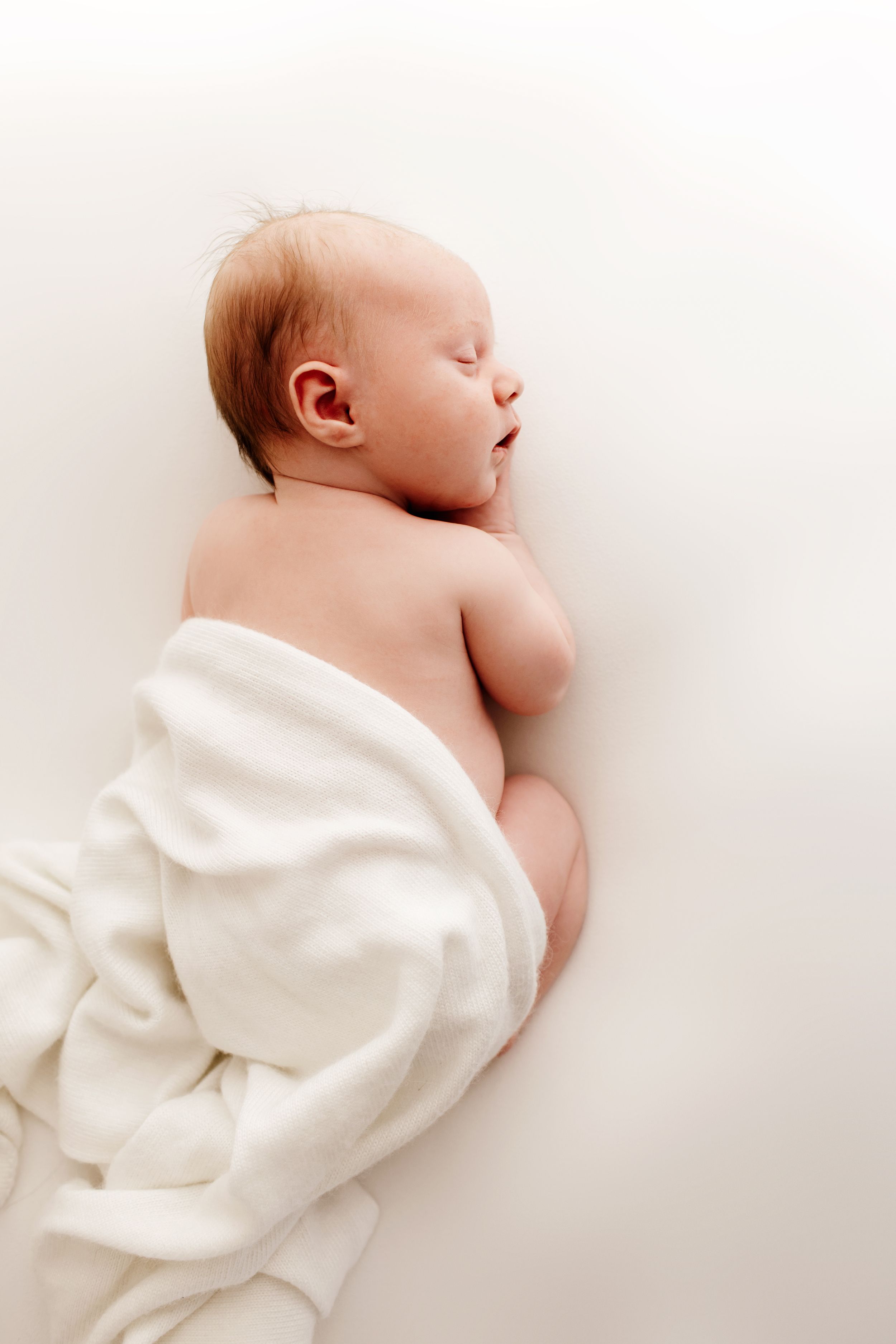 Peaceful newborn baby wrapped in white blanket resting on light background.