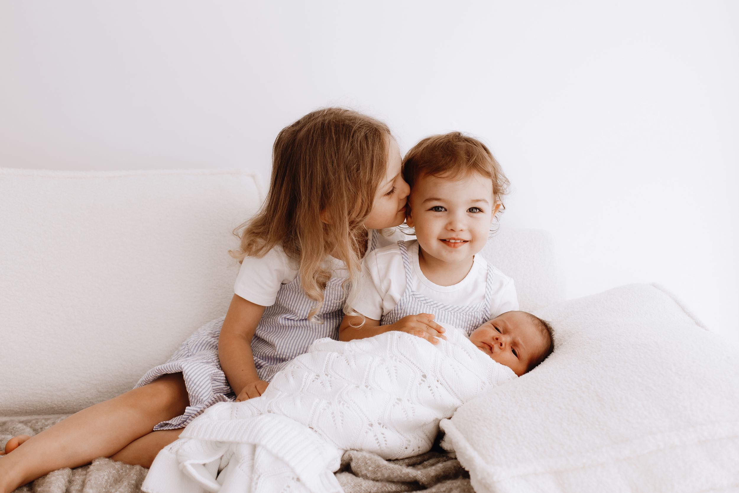 Candid family moments on white bedding showing siblings meeting new baby.