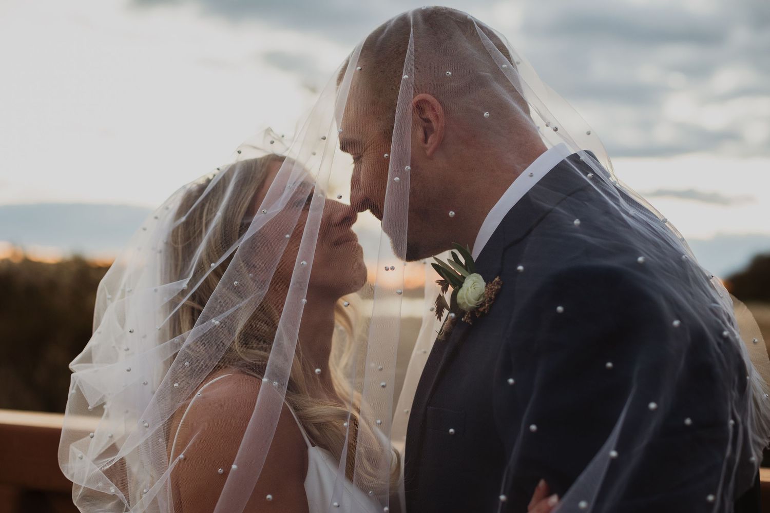 A romantic wedding couple embraces in a close-up shot with a veil flowing in the wind at sunset.