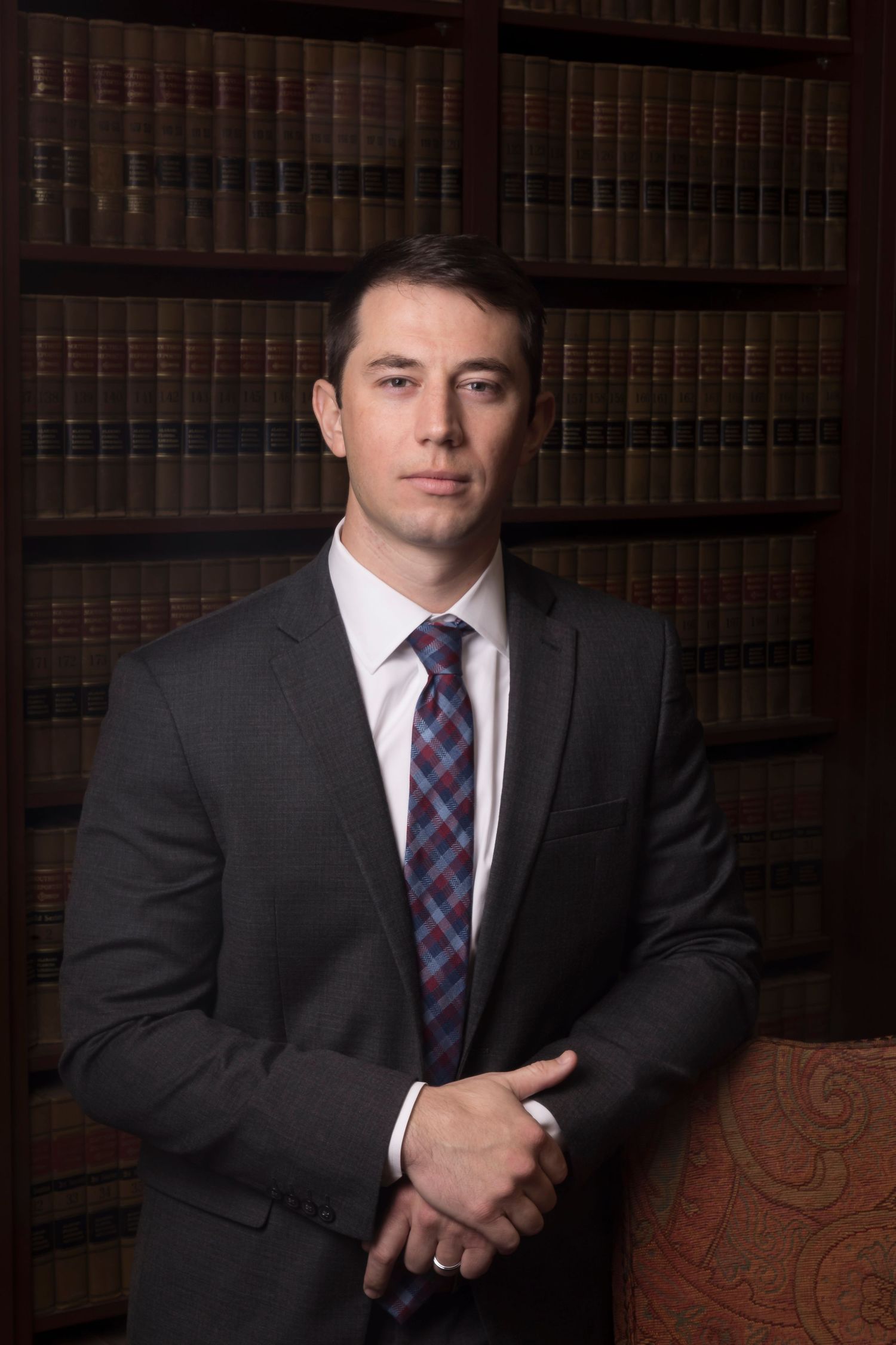 Professional headshot of a businessman in a dark suit and plaid tie standing in front of law library shelves.