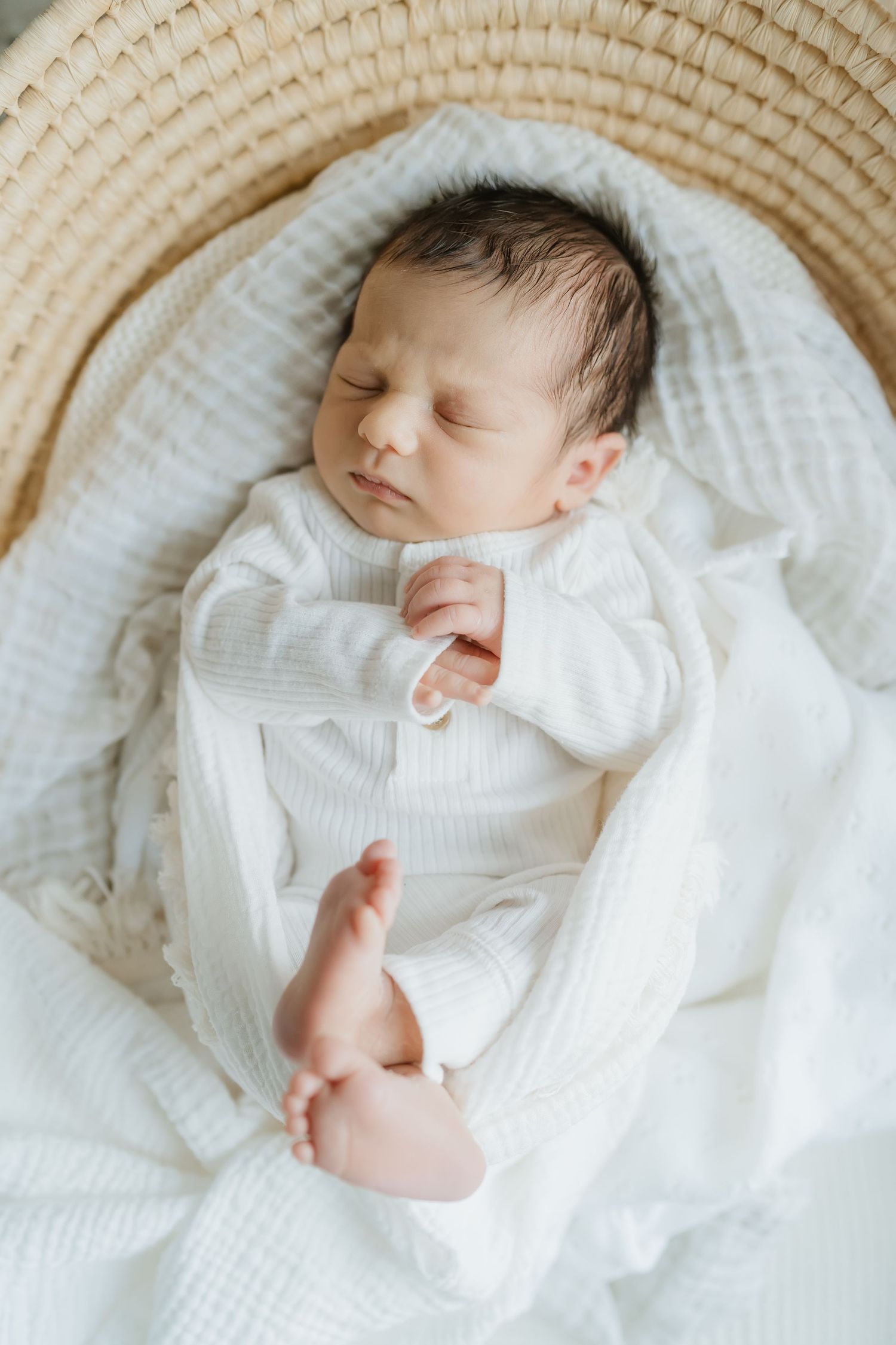 Sleeping newborn baby in white clothing nestled in cozy basket with soft white blankets.