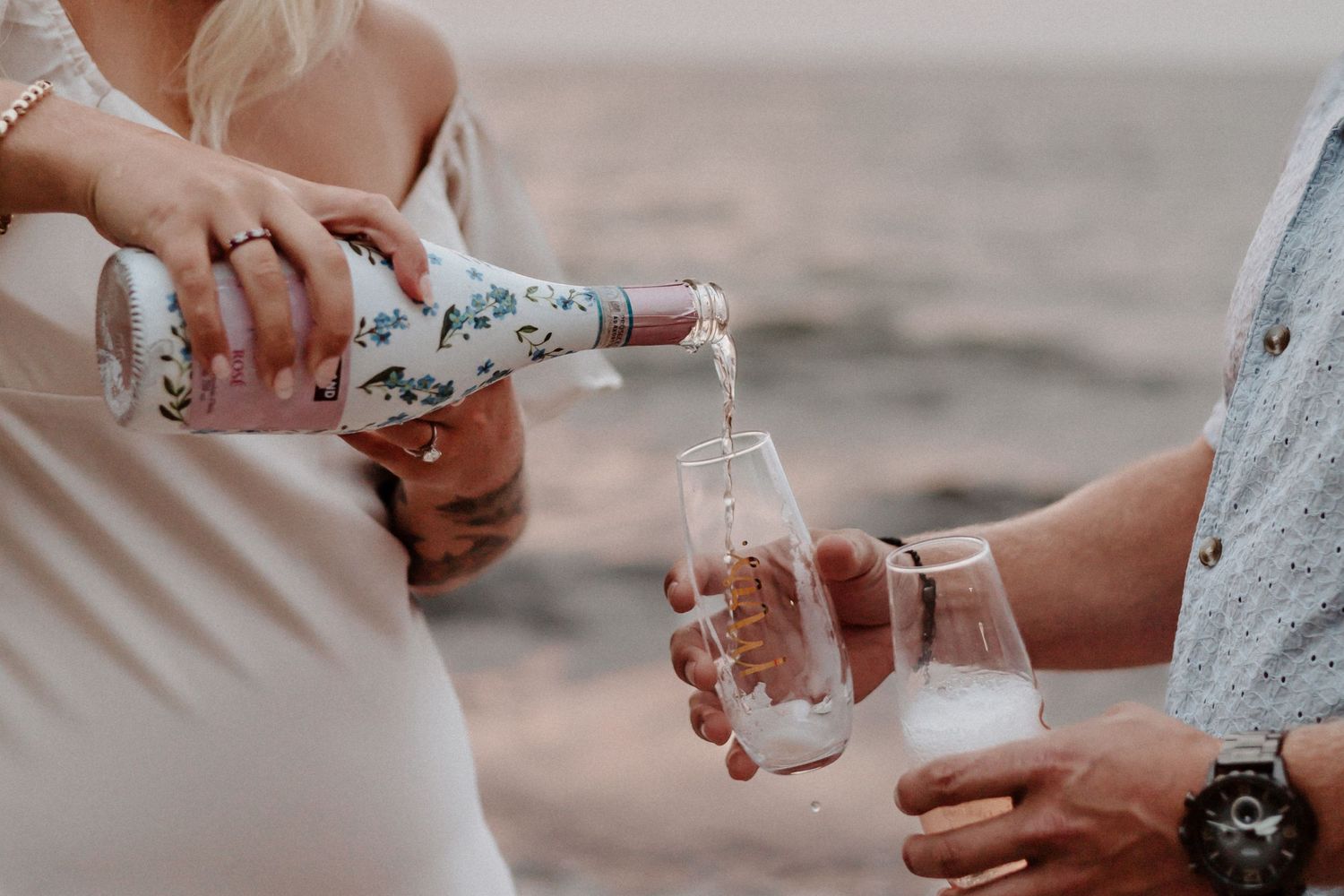 Hands pour champagne into glasses at a beach during sunset.