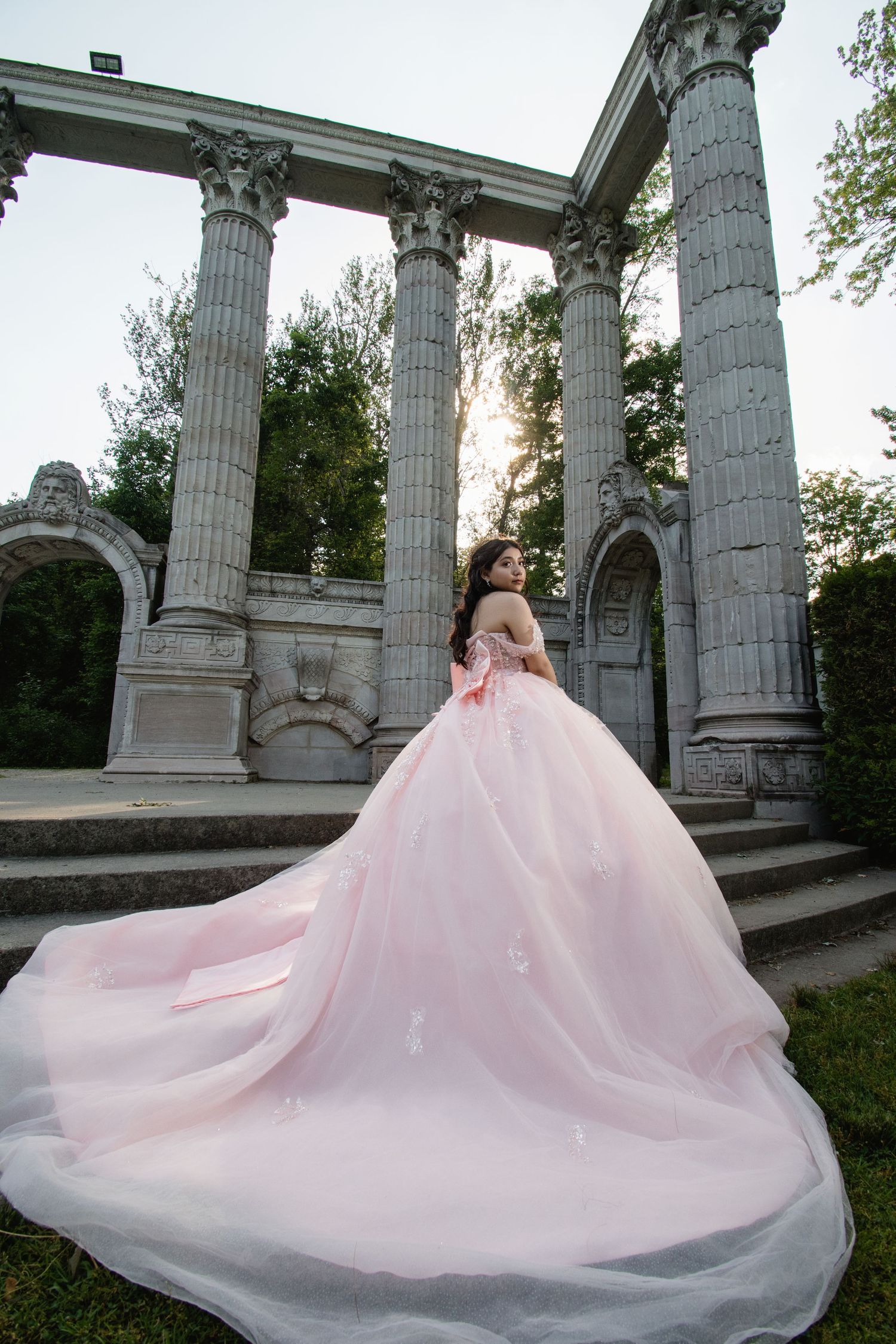 A bride in a voluminous pink ball gown stands before elegant stone columns in a garden at sunset. Toronto GTA