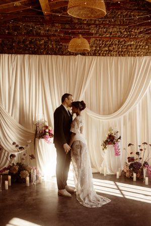 Silhouetted couple shares an intimate moment in front of flowing white drapes and floral arrangements in romantic venue lighting.