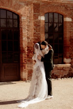 Intimate moment between newlyweds embracing at rustic brick venue with sunlit backdrop.