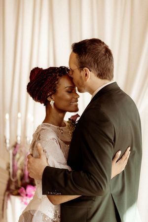 A couple in wedding attire share an intimate moment in front of white curtains with candles and flowers in the background.