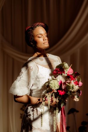 Artistic photo of a bride holding a romantic bouquet of pink and red flowers against a draped cream backdrop.