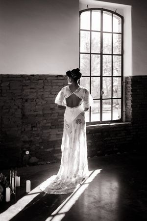 Dramatic silhouette of wedding dress hanging by window in rustic brick room with natural light streaming in.