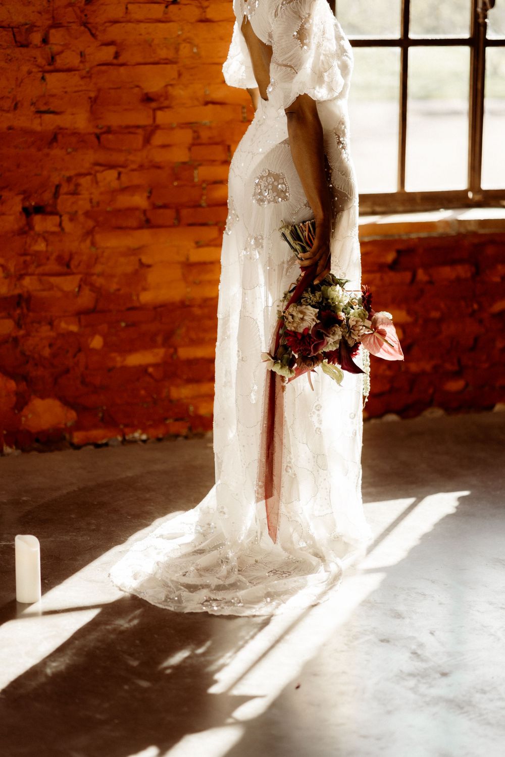 A bride in an elegant lace wedding dress holds a bouquet while standing in warm sunlight against a brick wall.