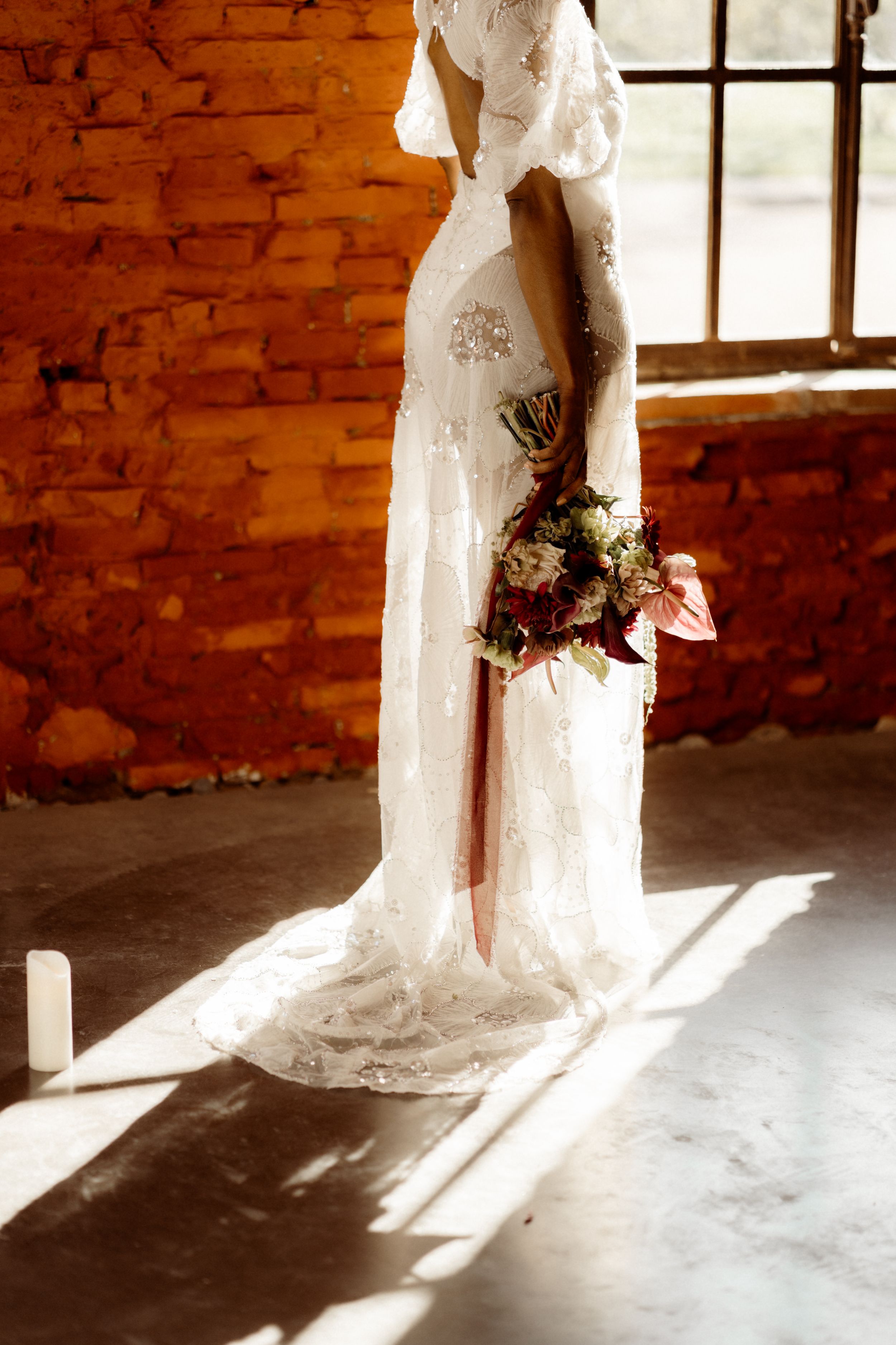 A bride in an elegant lace wedding dress holds a bouquet while standing in warm sunlight against a brick wall.