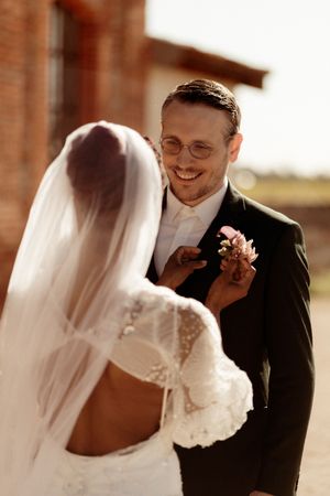 A groom in glasses beams with joy as he sees his bride in her lace wedding dress and veil.
