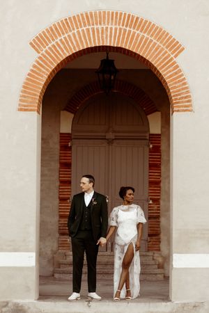 Stylish couple poses in a vintage brick archway, bride wearing elegant white dress with slit and groom in classic black suit.