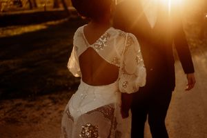 Romantic sunset wedding photo showing backs of couple with bride in lace backless dress walking on dirt path.