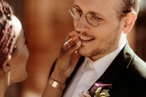 A close-up wedding moment shows a tender touch between a couple wearing formal attire with glasses and a floral boutonniere.