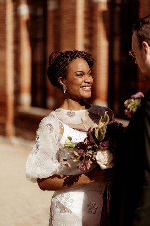 A romantic moment between newlyweds holding burgundy and cream floral bouquets in warm sunlight.